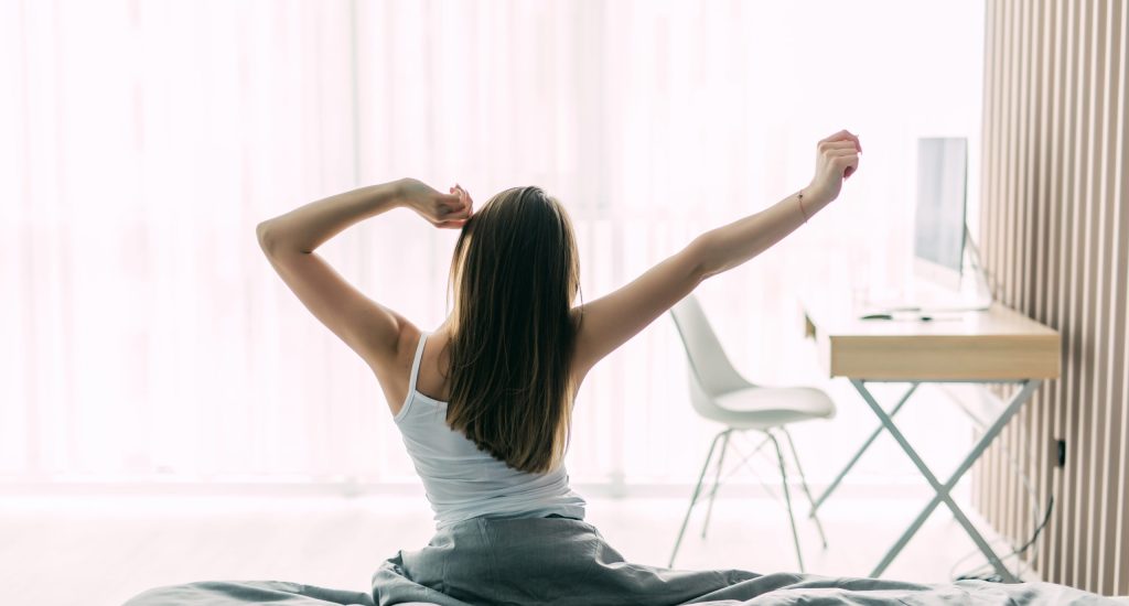 Back view of young woman stretching on unmade bed after waking up and looking at city view in the window. Motivation concept